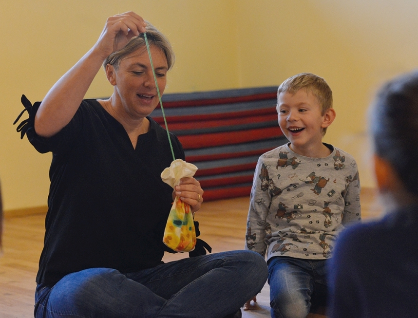 Eine Kindergartenpädagogin hält ein bemaltes Baumwollsäckchen in der Hand. Daraus zieht sie einen langen Faden. Neben ihr sitzt ein Bub, beobachtet den Faden und lacht fröhlich.