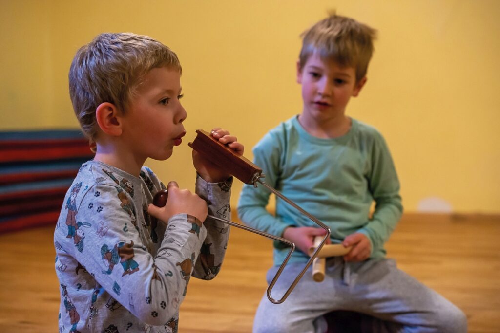 Zwei Buben sitzen am Boden und probieren Musikinstrumente aus. Der linke hält ein Schlaginstrument in den Händen und bläst hinein. Der rechte Bub hält Klangstäbe.