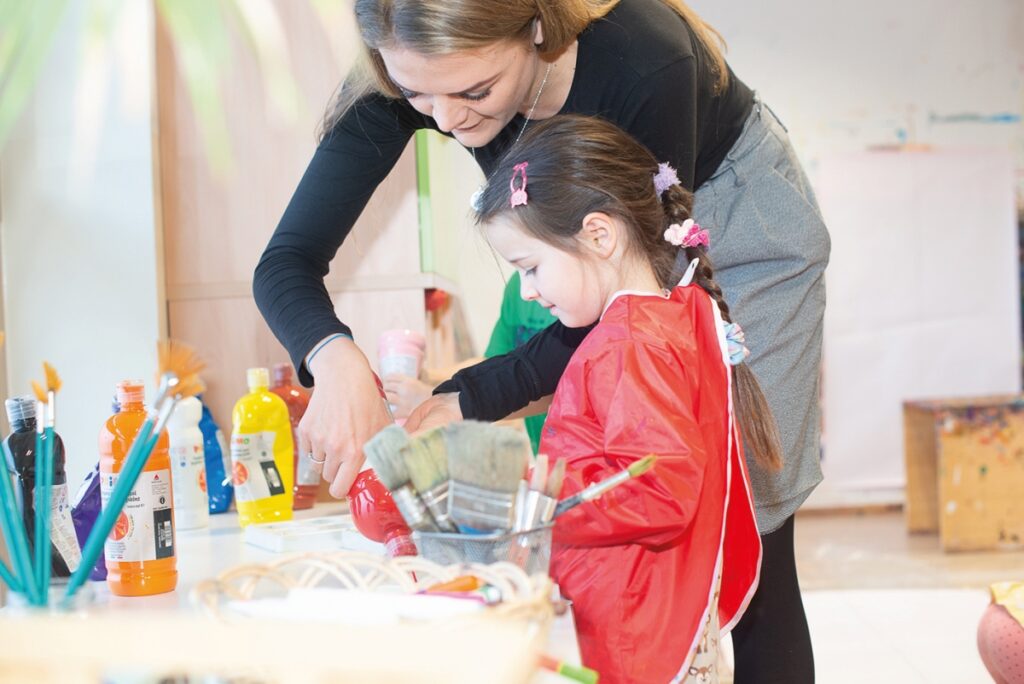 An einem Tisch mit vielen bunten Farbflaschen und Pinseln steht ein kleines Mädchen mit Malerschürze. Eine Kindergartenpädagogin hilft ihr beim Abfüllen der roten Farbe.