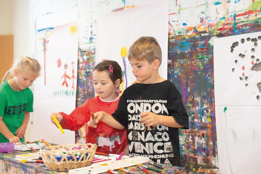 Drei Kinder stehen um einen kleinen Tisch mit Malutensilien. Hinter ihnen an der Wand hängen bereits halbfertige Kunstwerke.